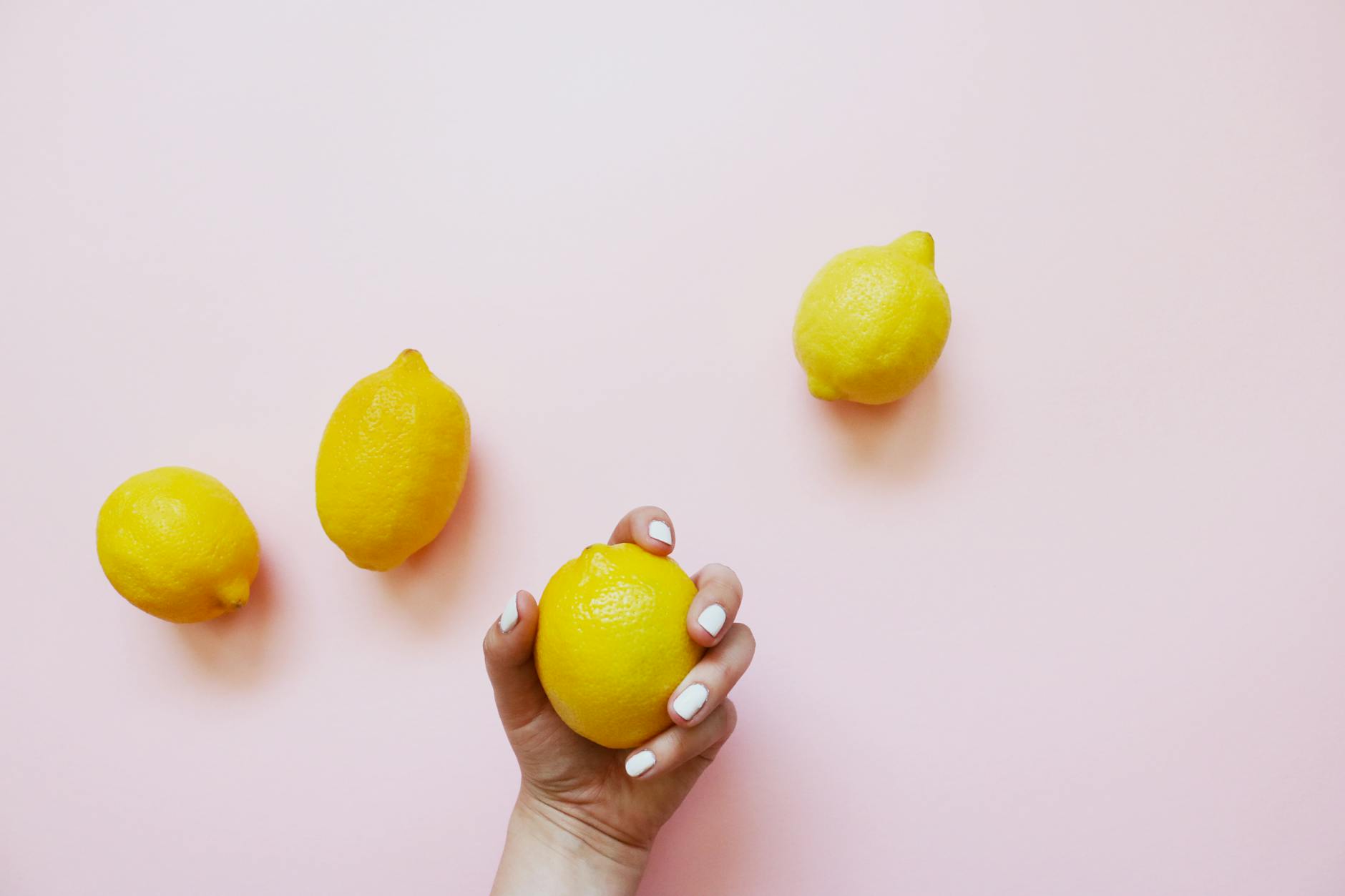 A hand with white nails holding a bright yellow lemon on a soft pink background, surrounded by additional lemons.